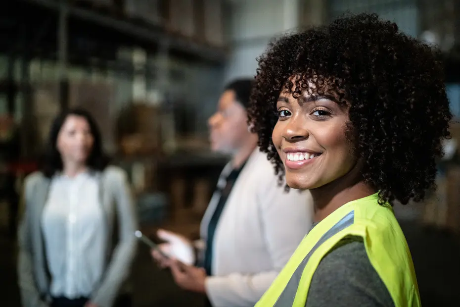 A smiling woman with curly hair wears a bright yellow safety vest in a warehouse setting. Two other women are blurred in the background, engaged in conversation. The focus is on the woman in the foreground, who exudes confidence and friendliness.