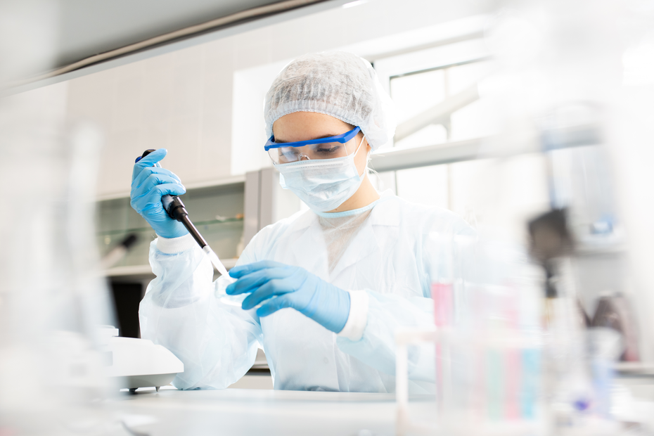A laboratory technician in protective gear, including gloves, a lab coat, a face mask, and goggles, is using a pipette to handle a liquid sample on a lab bench surrounded by laboratory equipment.