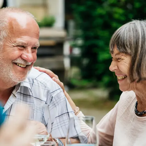 A joyful elderly couple shares a warm, intimate moment, smiling and laughing together at a gathering. Their expressions convey happiness and connection, embodying a sense of companionship and celebration.