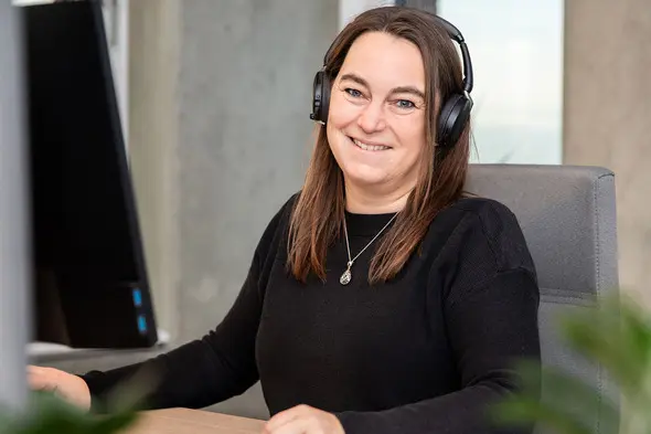 A woman wearing headphones smiles while seated at a desk with a computer monitor in the background. She has long brown hair and is wearing a black sweater. More foliage can be seen in the lower part of the image.