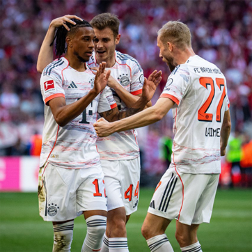 Three football players in white and orange uniforms from Bayern Munich celebrate a successful play. One player is joyfully clasping the shoulders of another, while the third extends a hand, rejoicing together in a moment of victory on the field.