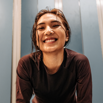 A smiling woman with wet hair leans forward, exuding joy and energy. She wears a dark athletic shirt and is set against a light blue background, suggesting a post-workout moment. Her expression reflects happiness and satisfaction.