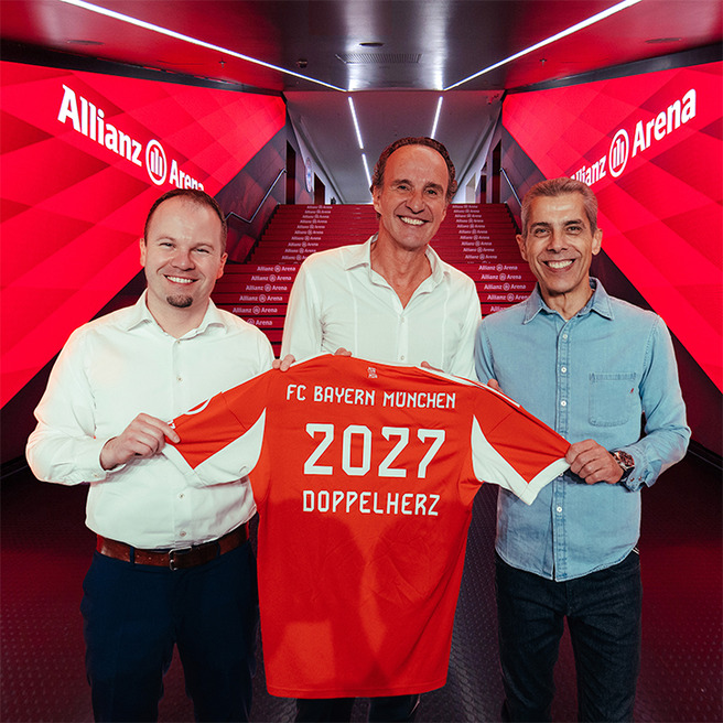 Three men are standing together, holding a Bayern Munich jersey displaying "2027" and "DOPPELHERZ." They are in a brightly lit corridor with red decorations and the "Allianz Arena" logo visible in the background. The atmosphere appears celebratory and significant.