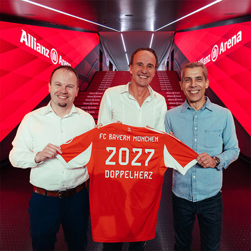 Three men are standing together, holding a Bayern Munich jersey displaying "2027" and "DOPPELHERZ." They are in a brightly lit corridor with red decorations and the "Allianz Arena" logo visible in the background. The atmosphere appears celebratory and significant.