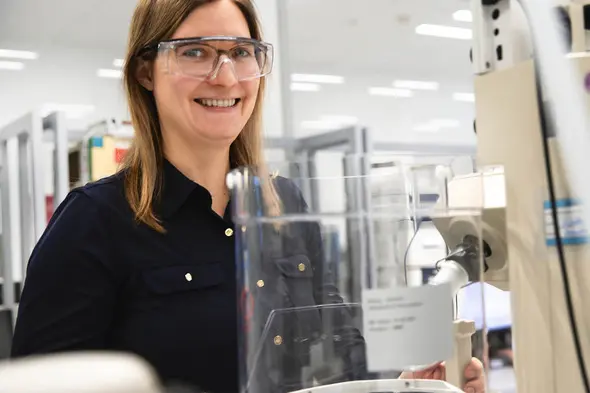 A smiling woman wearing safety glasses stands beside laboratory equipment. She is dressed in a dark shirt and appears engaged in her work environment, which features a bright, modern laboratory setting.