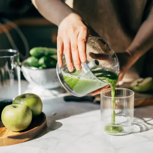 A person pours green juice from a blender into a clear glass. Fresh fruits, including green apples and cucumbers, are visible on a countertop, suggesting the preparation of a healthy beverage. Natural light illuminates the scene, highlighting the freshness of the ingredients.