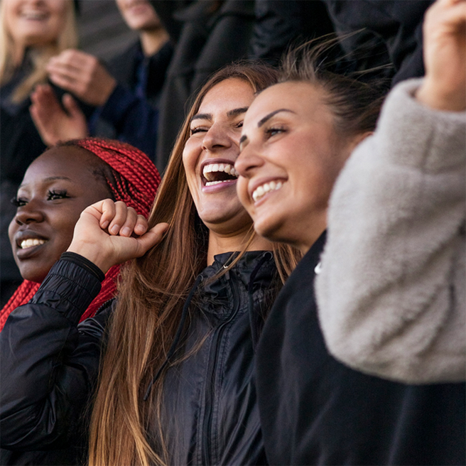 Three women are smiling and cheering enthusiastically, surrounded by others in a crowd. They are dressed in casual jackets, with one woman having long, red braids. The atmosphere conveys joy and excitement, likely in response to an event or performance.